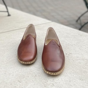 May include: A pair of brown leather slip-on shoes. The shoes have a rounded toe and a stitched sole. The leather has a polished finish. The shoes are displayed on a light-colored surface.