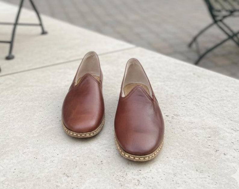 May include: A pair of brown leather slip-on shoes. The shoes have a rounded toe and a stitched sole. The leather has a polished finish. The shoes are displayed on a light-colored surface.