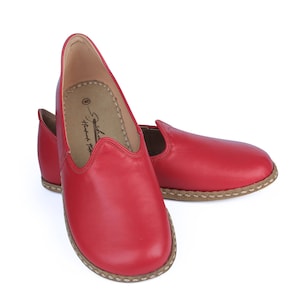 Puede incluir: Un par de zapatos sin cordones de cuero rojo. Los zapatos tienen una puntera redondeada y una suela cosida de color marr&oacute;n claro. El interior del zapato es de color marr&oacute;n claro. Los zapatos est&aacute;n sobre un fondo blanco.