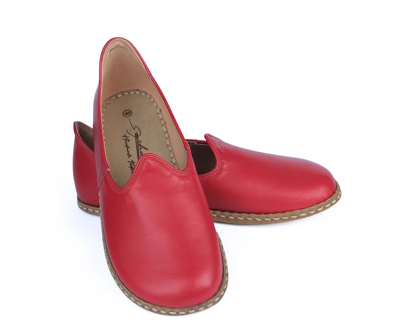 Puede incluir: Un par de zapatos sin cordones de cuero rojo. Los zapatos tienen una puntera redondeada y una suela cosida de color marr&oacute;n claro. El interior del zapato es de color marr&oacute;n claro. Los zapatos est&aacute;n sobre un fondo blanco.