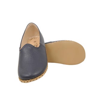 May include: A pair of navy blue slip-on shoes with a textured leather-like upper, a rounded toe, and a light tan sole. The shoes feature visible light-colored stitching along the edge of the sole and a beige interior lining with a small logo.