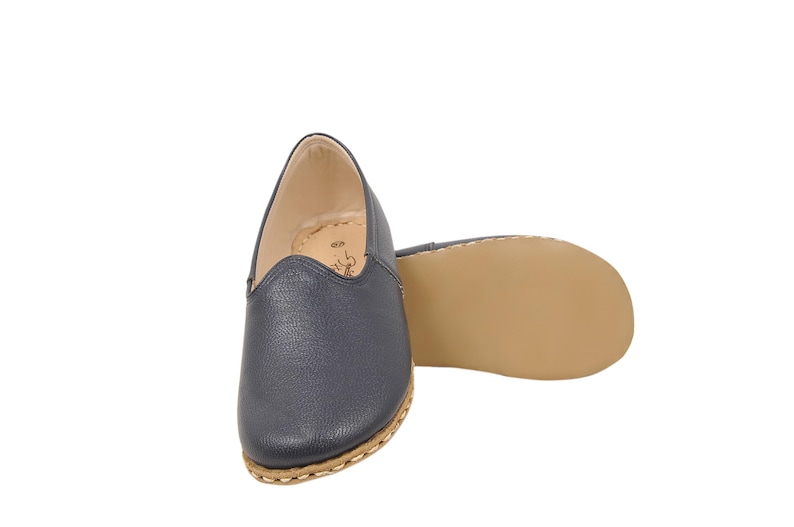 May include: A pair of navy blue slip-on shoes with a textured leather-like upper, a rounded toe, and a light tan sole. The shoes feature visible light-colored stitching along the edge of the sole and a beige interior lining with a small logo.