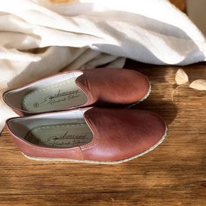 May include: A pair of brown slip-on shoes with a rounded toe and a visible stitched sole edge. The interior insoles feature the text "charooy Handmade Footwear" printed in black script. The shoes have a smooth, solid-colored exterior finish.