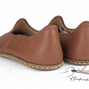 May include: A pair of brown leather slip-on shoes with a stitched seam around the sole. The shoes are handmade and feature a simple, elegant design.