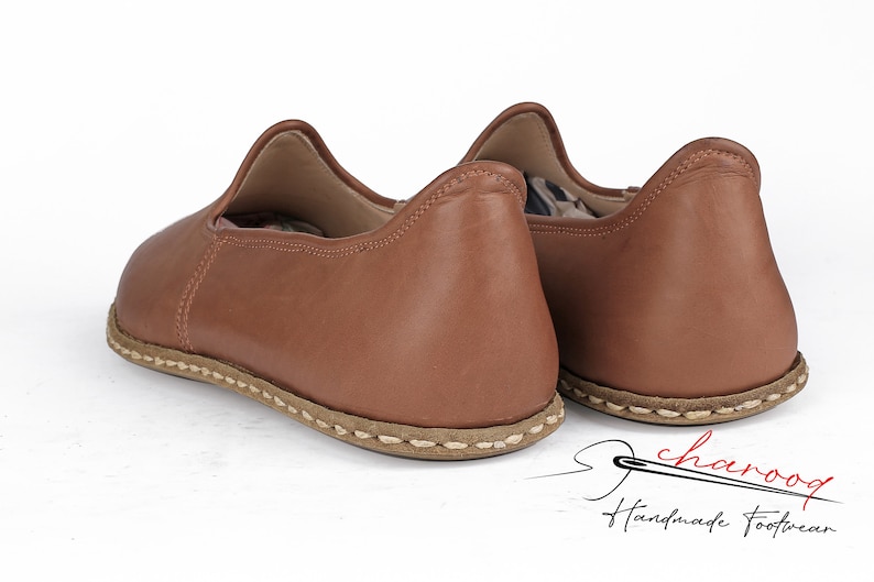 May include: A pair of brown leather slip-on shoes with a stitched seam around the sole. The shoes are handmade and feature a simple, elegant design.
