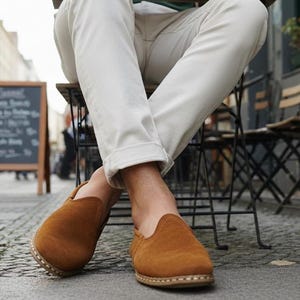K&ouml;nnte beinhalten: Nahaufnahme von braunen Wildleder-Slippern mit runder Spitze. Die Schuhe werden von einer Person getragen, die in einem Stra&szlig;encaf&eacute; sitzt. Die Person tr&auml;gt eine wei&szlig;e Hose. Eine Tafel mit Speisekarte ist im Hintergrund.