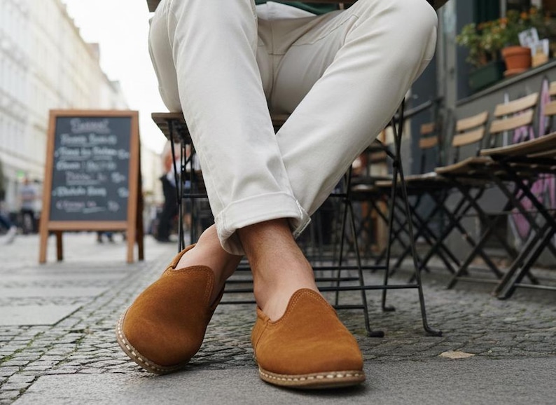 K&ouml;nnte beinhalten: Nahaufnahme von braunen Wildleder-Slippern mit runder Spitze. Die Schuhe werden von einer Person getragen, die in einem Stra&szlig;encaf&eacute; sitzt. Die Person tr&auml;gt eine wei&szlig;e Hose. Eine Tafel mit Speisekarte ist im Hintergrund.