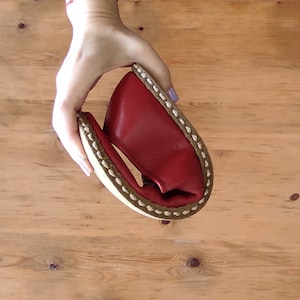 Puede incluir: Un zapato de cuero rojo se sostiene en la mano, mostrando su flexibilidad. El zapato tiene una suela blanca con un borde cosido marr&oacute;n. El fondo es una superficie de madera marr&oacute;n claro.