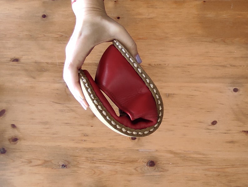 Puede incluir: Un zapato de cuero rojo se sostiene en la mano, mostrando su flexibilidad. El zapato tiene una suela blanca con un borde cosido marr&oacute;n. El fondo es una superficie de madera marr&oacute;n claro.