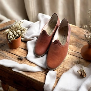 May include: A pair of brown leather slip-on shoes with a rounded toe and a stitched sole. The shoes are displayed on a rustic wooden table with a small terracotta pot of flowers and a decorative vase.
