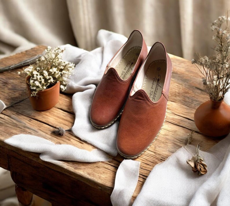 May include: A pair of brown leather slip-on shoes with a rounded toe and a stitched sole. The shoes are displayed on a rustic wooden table with a small terracotta pot of flowers and a decorative vase.