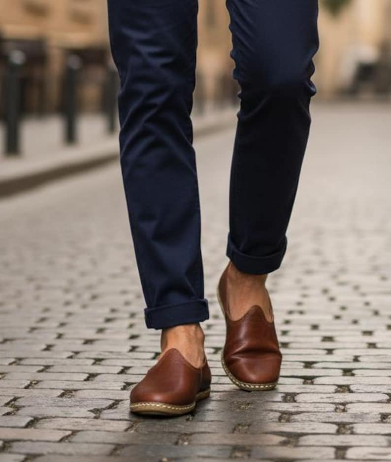 May include: Brown leather slip-on shoes with a rounded toe and a stitched sole. The shoes are worn with navy blue pants, cuffed at the ankle. The image is taken on a cobblestone street.