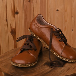 May include: Two pairs of brown leather lace-up shoes with brown laces. The shoes have a stitched seam around the sole and are sitting on a wooden surface.