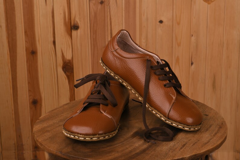 May include: Two pairs of brown leather lace-up shoes with brown laces. The shoes have a stitched seam around the sole and are sitting on a wooden surface.