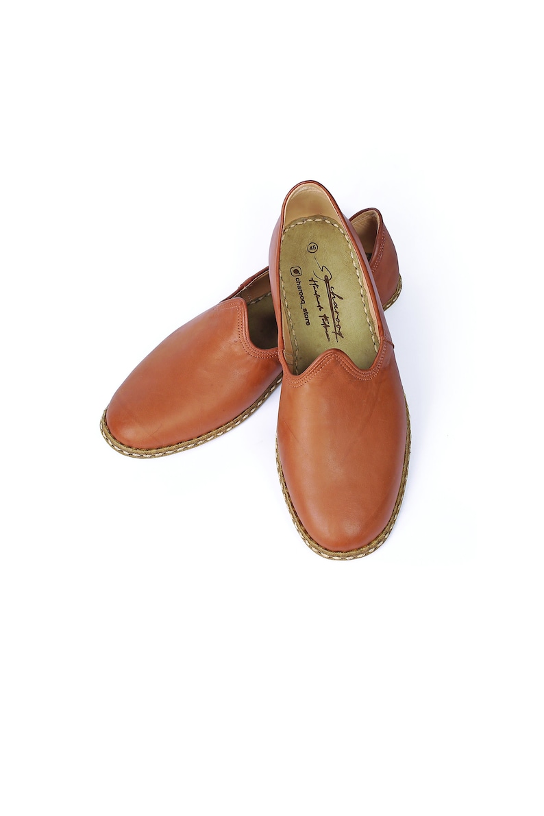 May include: A pair of brown leather slip-on shoes with a stitched edge and a brown leather sole. The shoes have a pointed toe and a simple design. The brand name "Leatherfoot Handmade Footwear" is printed on the insole.