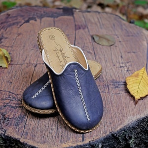 May include: A pair of dark blue leather slippers with white stitching and a brown leather sole. The slippers are on a wooden surface with some fallen leaves.