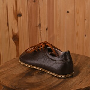 May include: A brown leather shoe with a stitched sole and orange laces. The shoe is sitting on a wooden surface.