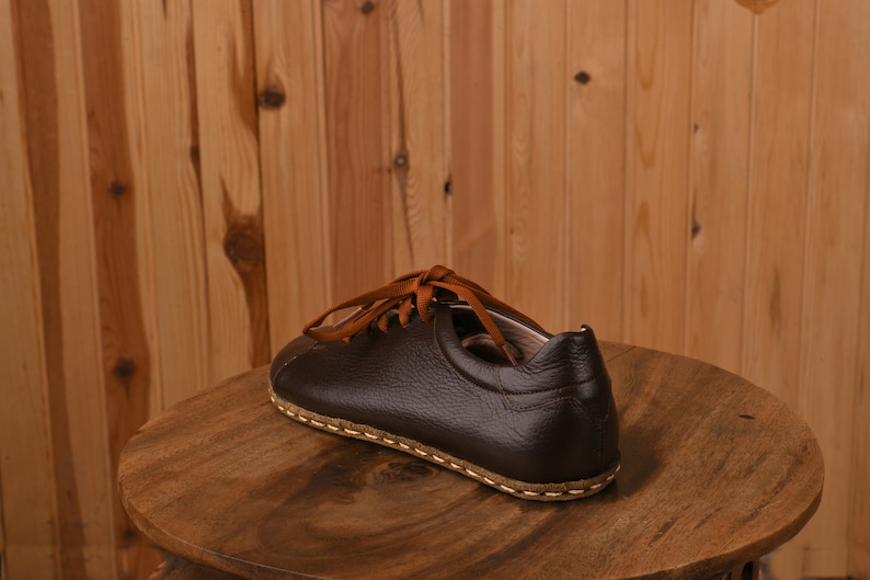 May include: A brown leather shoe with a stitched sole and orange laces. The shoe is sitting on a wooden surface.