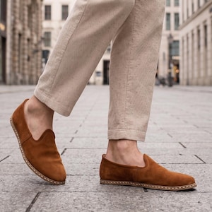 K&ouml;nnte beinhalten: Braune Wildleder-Slipper mit runder Spitze und Ziern&auml;hten. Die Schuhe haben einen warmen, erdigen Farbton, und die Sohlen scheinen aus einem hellen Material zu bestehen. Getragen mit beigefarbenen Cordhosen.