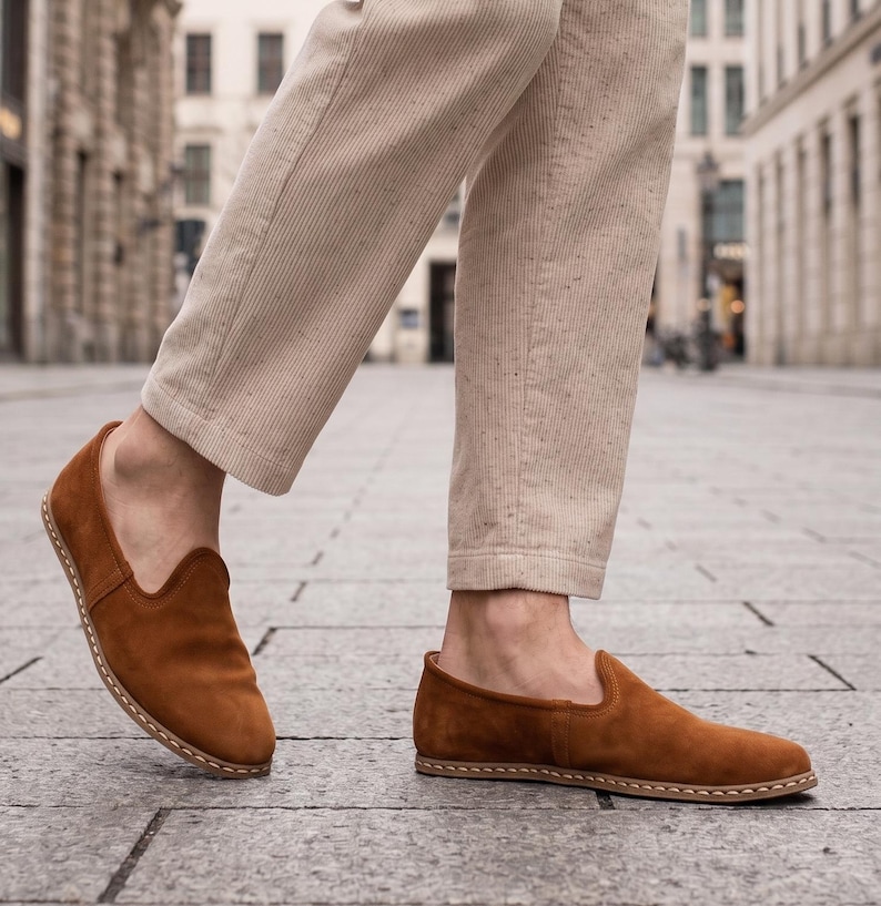 K&ouml;nnte beinhalten: Braune Wildleder-Slipper mit runder Spitze und Ziern&auml;hten. Die Schuhe haben einen warmen, erdigen Farbton, und die Sohlen scheinen aus einem hellen Material zu bestehen. Getragen mit beigefarbenen Cordhosen.