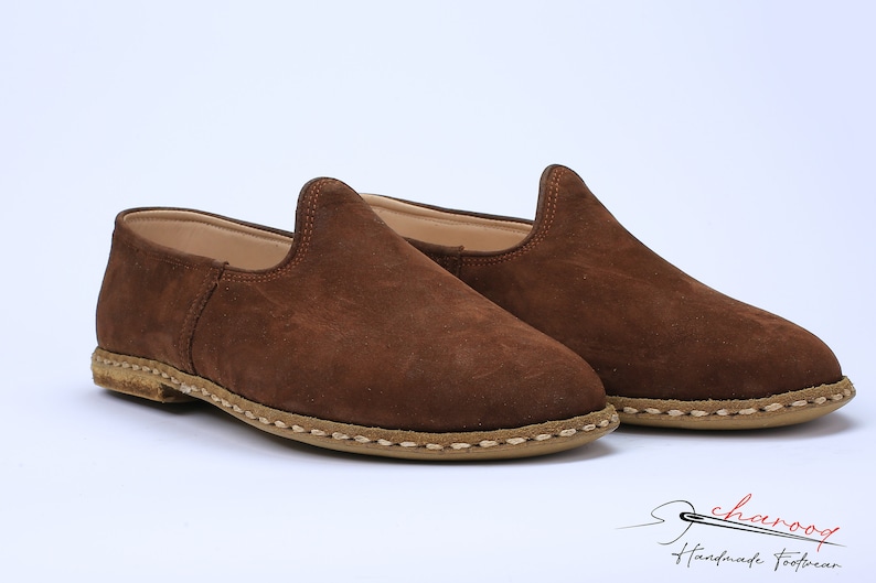 May include: A pair of brown suede slip-on shoes with a rounded toe, a high-cut vamp, and a tan sole featuring visible thick beige stitching around the perimeter. The logo for charooq Handmade Footwear is printed in the bottom right corner.