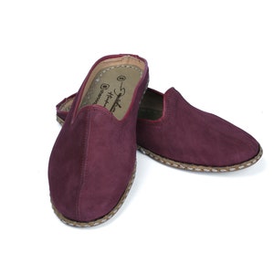 May include: A pair of burgundy suede slippers with a braided trim around the sole. The slippers have a pointed toe and a slip-on design.