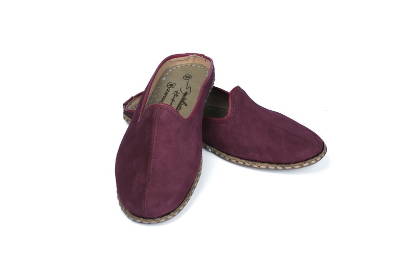 May include: A pair of burgundy suede slippers with a braided trim around the sole. The slippers have a pointed toe and a slip-on design.