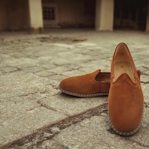 K&ouml;nnte beinhalten: Ein Paar braune Wildleder-Slipper. Die Schuhe haben eine runde Spitze und ein niedriges Profil mit sichtbaren N&auml;hten um die Sohle. Die Schuhe stehen auf einer strukturierten, grauen Steinoberfl&auml;che.