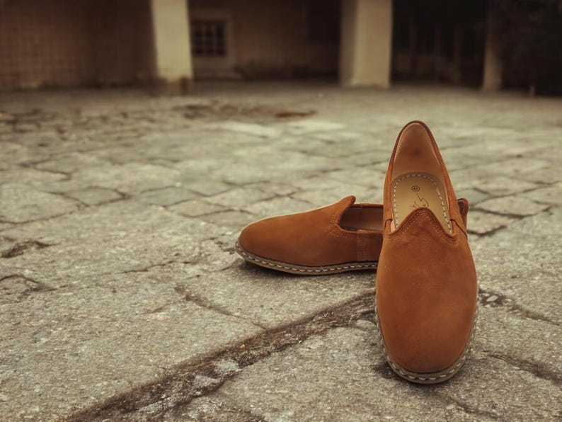 K&ouml;nnte beinhalten: Ein Paar braune Wildleder-Slipper. Die Schuhe haben eine runde Spitze und ein niedriges Profil mit sichtbaren N&auml;hten um die Sohle. Die Schuhe stehen auf einer strukturierten, grauen Steinoberfl&auml;che.
