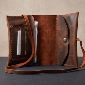 Puede incluir: Una bolsa de tabaco de cuero marrón con diseño de tres pliegues, con costuras visibles, cierre de botón a presión de metal y una correa de cuero integrada. El interior incluye un bolsillo con ranuras verticales para papel de fumar.