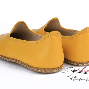 May include: A pair of yellow leather slip-on shoes with a stitched edge. The shoes are handmade and have a simple design.