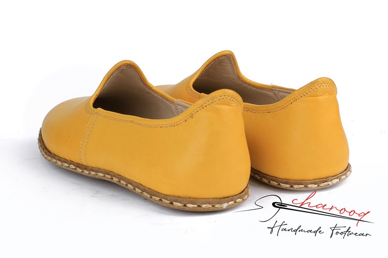 May include: A pair of yellow leather slip-on shoes with a stitched edge. The shoes are handmade and have a simple design.