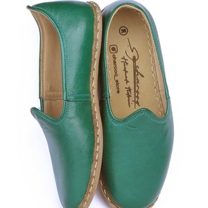 May include: A pair of green leather slippers with a brown leather sole and a stitched edge. The slippers have a pointed toe and a small flap at the top. The inside of the slippers is lined with a tan fabric. The slippers are labeled "Selam" and "Handmade Turkish Slippers" on the inside.