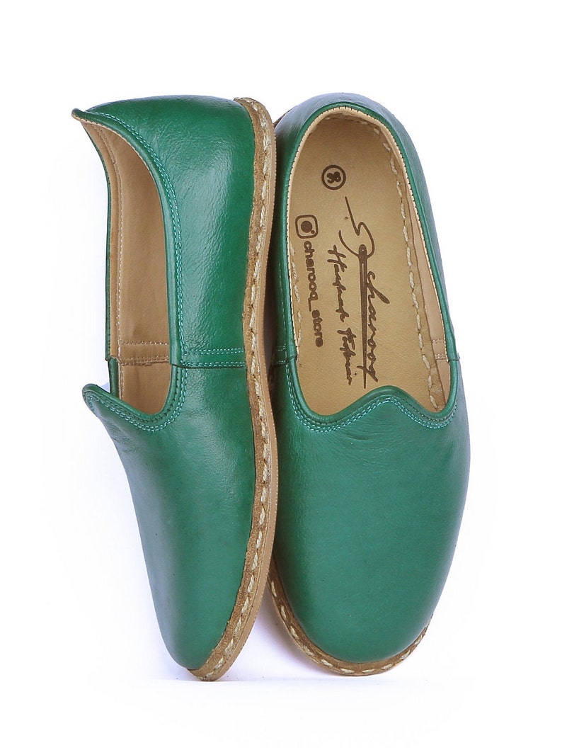 May include: A pair of green leather slippers with a brown leather sole and a stitched edge. The slippers have a pointed toe and a small flap at the top. The inside of the slippers is lined with a tan fabric. The slippers are labeled "Selam" and "Handmade Turkish Slippers" on the inside.