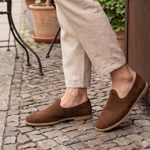 May include: A pair of brown suede slip-on shoes featuring a rounded toe, a low-cut topline, and a visible light-colored stitched welt detail along the sole. The shoes are worn with light beige trousers, showcasing a simple and clean design.