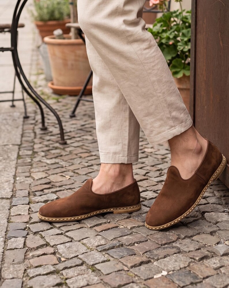 May include: A pair of brown suede slip-on shoes featuring a rounded toe, a low-cut topline, and a visible light-colored stitched welt detail along the sole. The shoes are worn with light beige trousers, showcasing a simple and clean design.