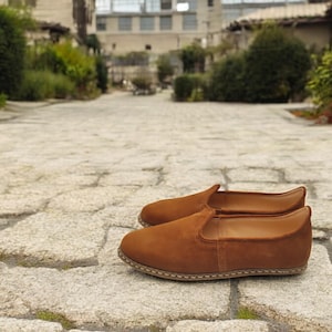 K&ouml;nnte beinhalten: Ein Paar braune Wildleder-Slipper. Die Schuhe haben eine runde Spitze und eine gen&auml;hte Sohle. Der Hintergrund ist ein Steinweg mit Gr&uuml;n und Geb&auml;uden in der Ferne. Die Schuhe haben einen warmen, erdigen Farbton.