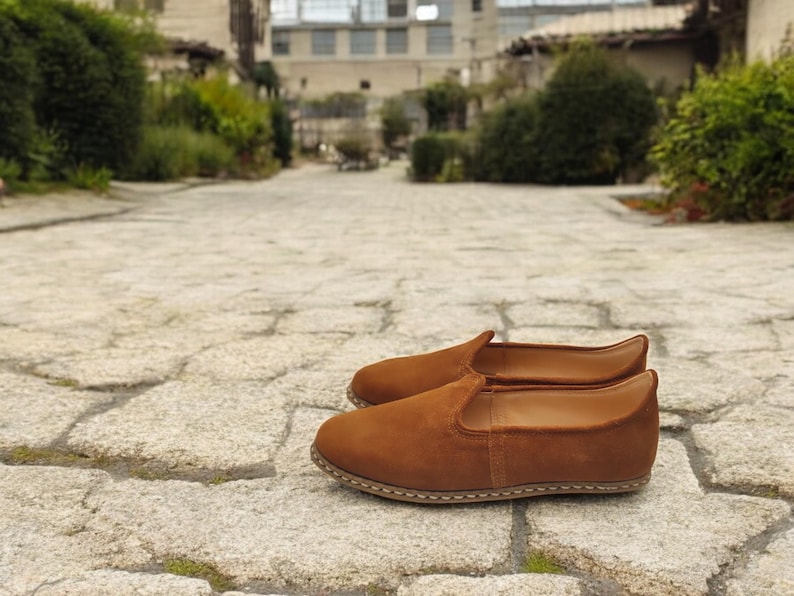 K&ouml;nnte beinhalten: Ein Paar braune Wildleder-Slipper. Die Schuhe haben eine runde Spitze und eine gen&auml;hte Sohle. Der Hintergrund ist ein Steinweg mit Gr&uuml;n und Geb&auml;uden in der Ferne. Die Schuhe haben einen warmen, erdigen Farbton.