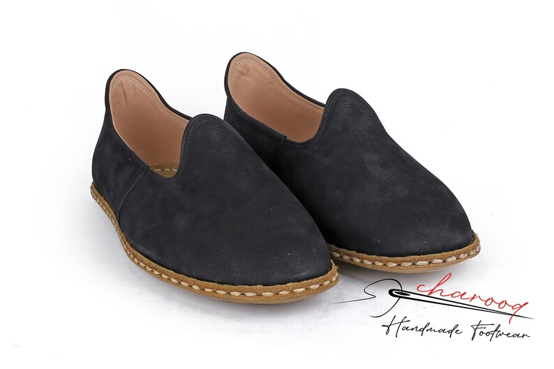 Puede incluir: Un par de zapatos negros de cuero sin cordones con un ribete cosido marr&oacute;n alrededor de la suela. Los zapatos son hechos a mano y tienen un aspecto casual y c&oacute;modo.