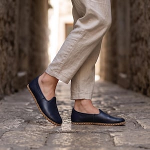 May include: A pair of navy blue slip-on shoes with a smooth finish and visible light-colored stitching along the sole edge. The shoes feature a low-cut profile and a rounded toe design, paired with light beige trousers that reach above the ankles.