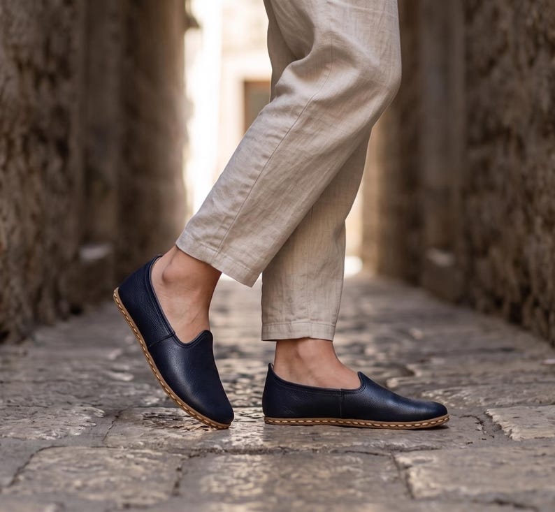 May include: A pair of navy blue slip-on shoes with a smooth finish and visible light-colored stitching along the sole edge. The shoes feature a low-cut profile and a rounded toe design, paired with light beige trousers that reach above the ankles.