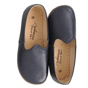 May include: A pair of navy blue slip-on shoes with a textured leather exterior. The interior features a light tan lining with the text "Scharooy Handmade Footwear" and the number "37" printed in black. The shoes have a rounded toe design.