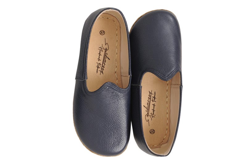May include: A pair of navy blue slip-on shoes with a textured leather exterior. The interior features a light tan lining with the text "Scharooy Handmade Footwear" and the number "37" printed in black. The shoes have a rounded toe design.