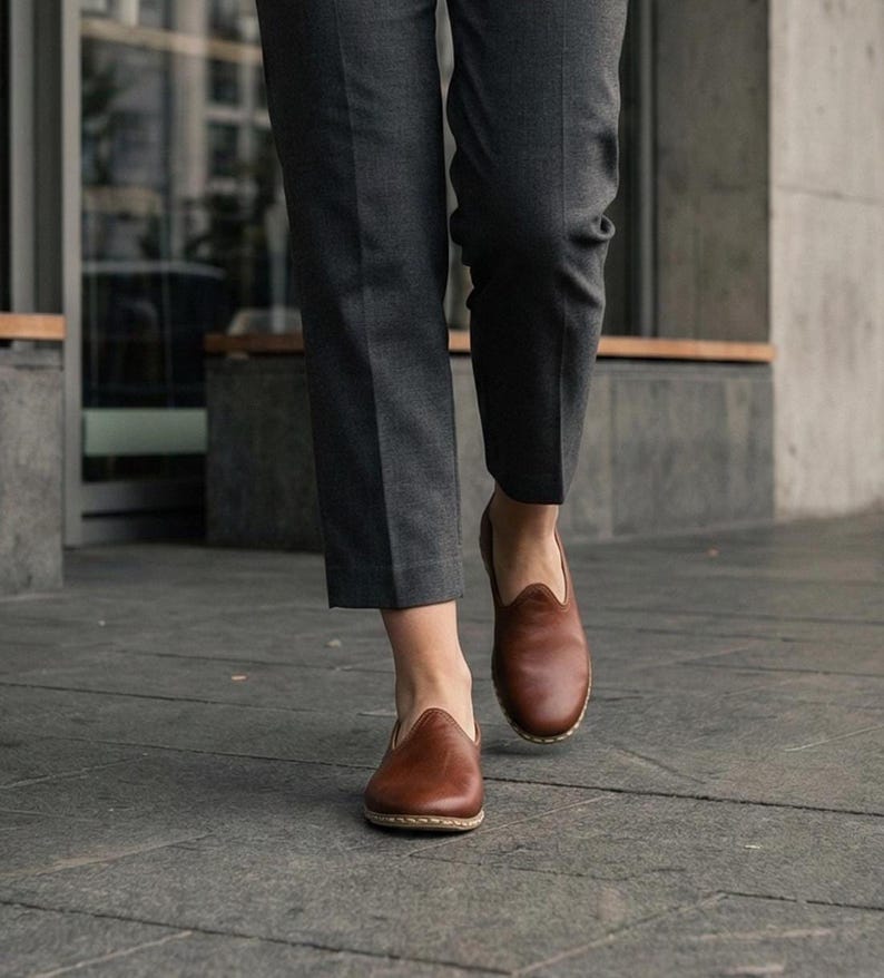 May include: A pair of brown leather slip-on shoes with a rounded toe and visible light-colored stitching along the sole, worn with dark gray tailored trousers that end above the ankle, showcasing a clean and simple design for everyday footwear.