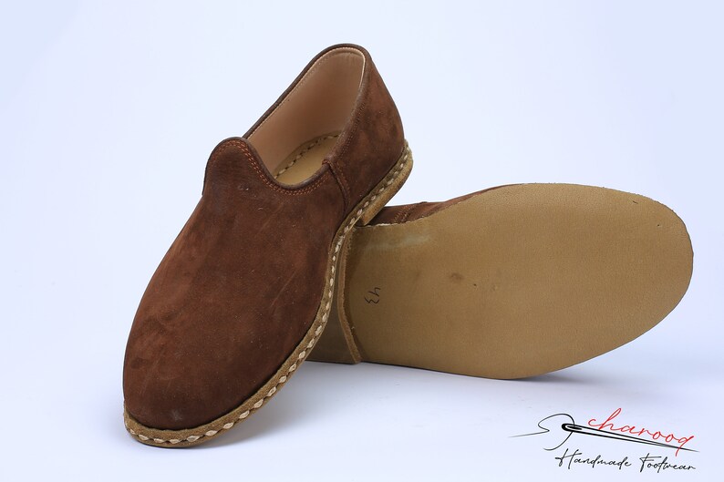 May include: A pair of brown suede slip-on shoes with a rounded toe and a light-colored stitched welt along the base. The sole is flat and tan, featuring the number 43 stamped on the bottom. The logo charooq Handmade Footwear is visible.