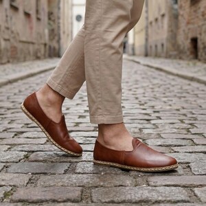 May include: Brown leather slip-on shoes with a woven sole. The shoes are a rich, warm brown color and appear to be made of high-quality leather. The shoes are being worn with tan pants. The shoes are a casual style.