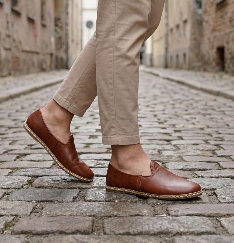 May include: Brown leather slip-on shoes with a woven sole. The shoes are a rich, warm brown color and appear to be made of high-quality leather. The shoes are being worn with tan pants. The shoes are a casual style.