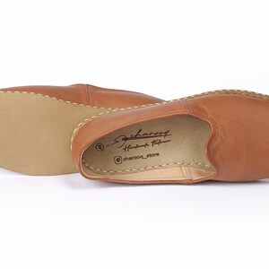 May include: A pair of brown leather slip-on shoes with a stitched edge. The shoes have a simple design and are made of soft leather. The insole is labeled "charoof Handmade Footwear" and "@charoof_store".