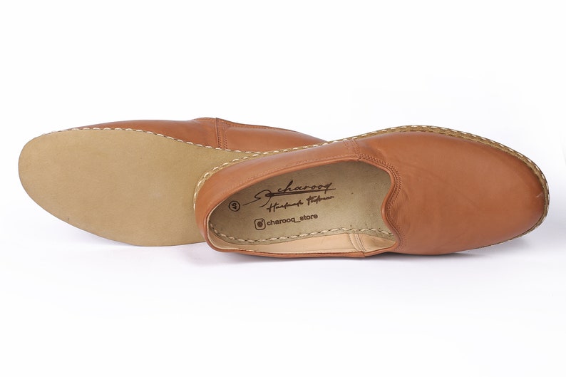 May include: A pair of brown leather slip-on shoes with a stitched edge. The shoes have a simple design and are made of soft leather. The insole is labeled "charoof Handmade Footwear" and "@charoof_store".