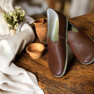 May include: A pair of brown leather slip-on shoes with white stitching around the edges. The shoes are on a wooden surface.